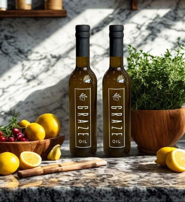 Two bottles of Graze Oil on a kitchen counter with lemons, herbs, and a glass of red wine.