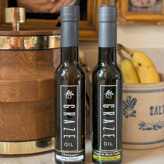 Two bottles of Graze Oil on a surface with a wooden barrel and bananas in the background.