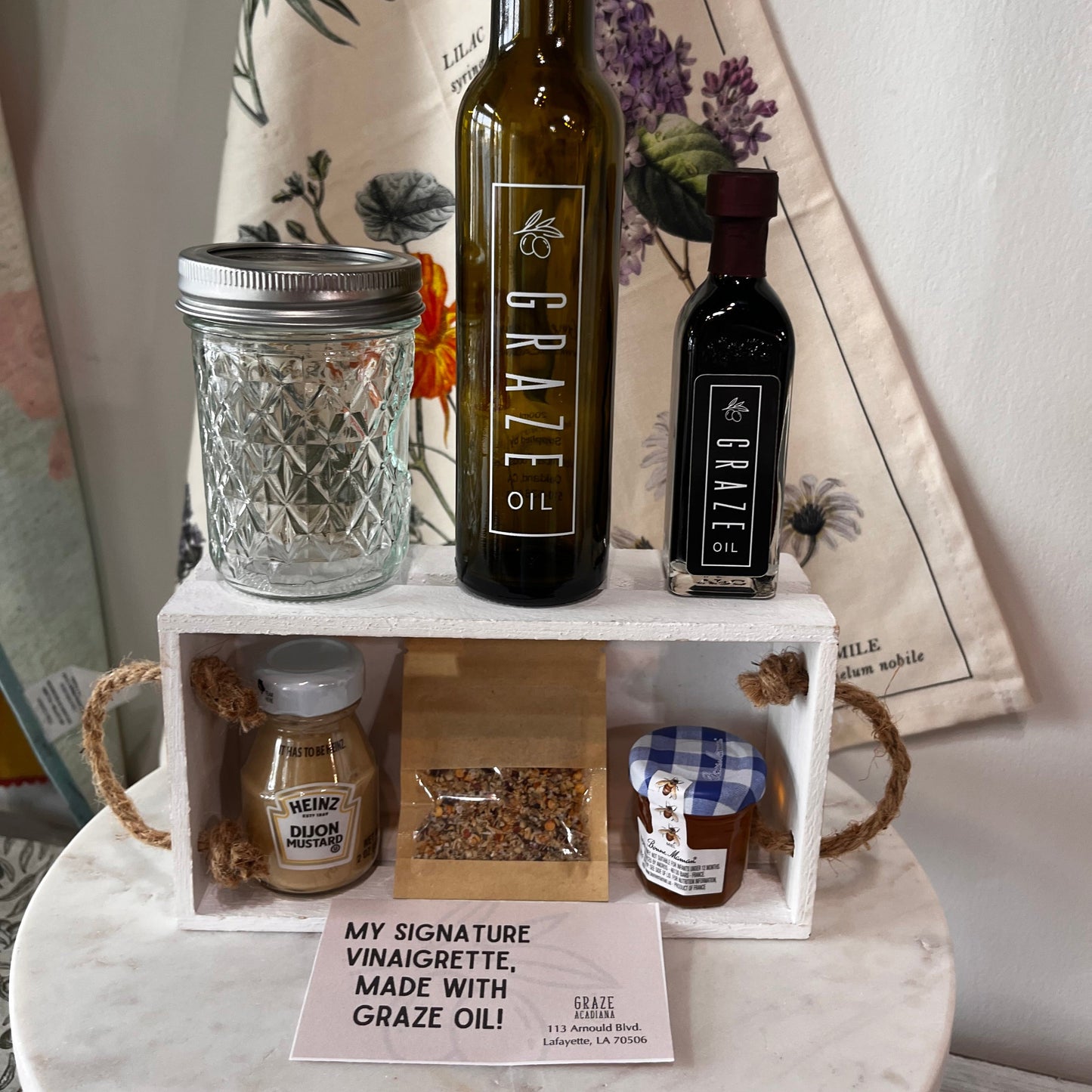 A gift set of gourmet food items displayed on a white wooden crate with rope handles, sitting on a round white marble side table. The set includes a bottle of Graze Oil, a smaller bottle of dark balsamic vinegar, a glass mason jar, a jar of honey, a small bottle of mustard, and a bag of seeds, with a sign reading 'MY SIGNATURE VINAIGRETTE, MADE WITH GRAZE OIL!'.
