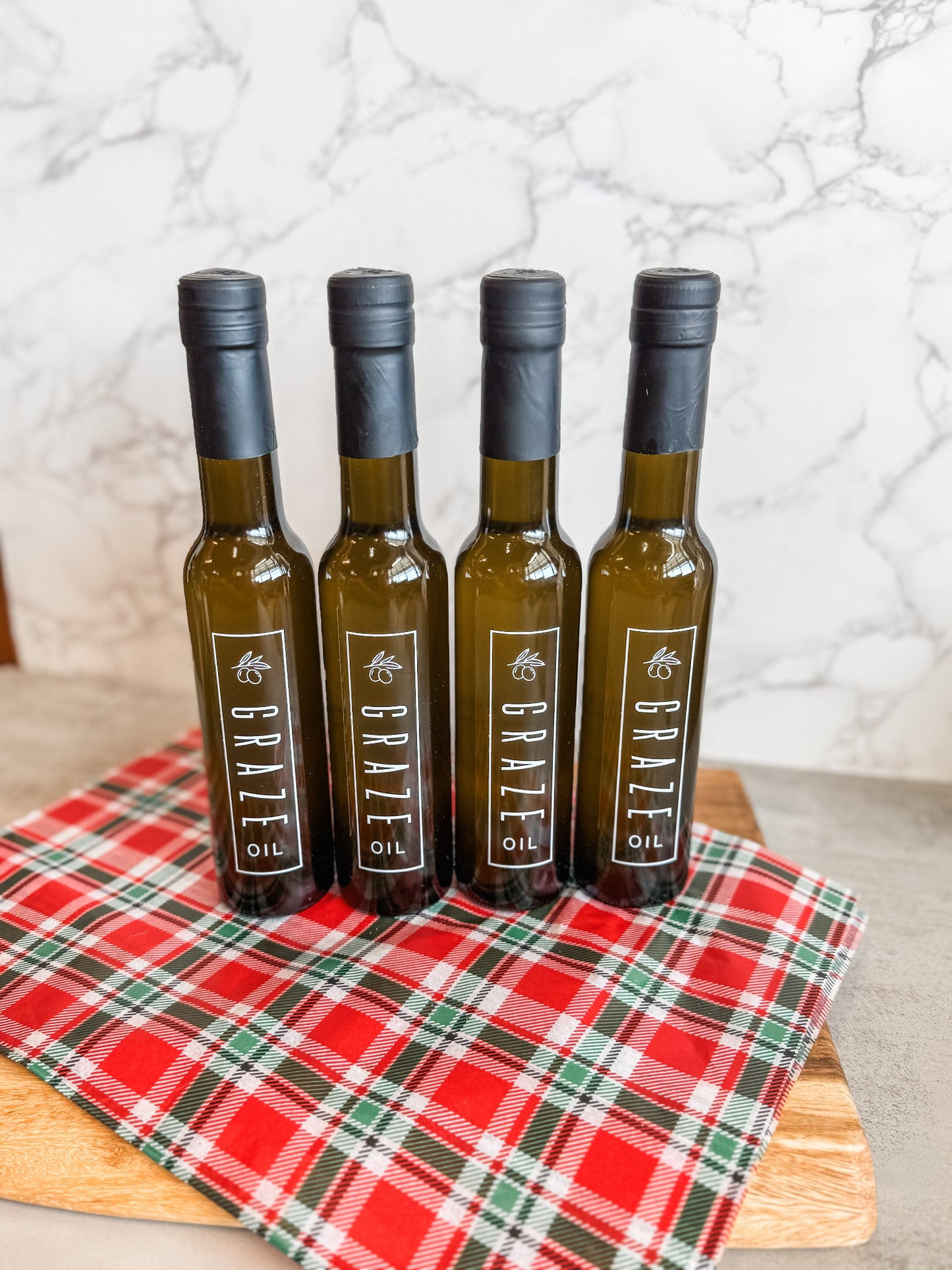 Four bottles of Graze Oil olive oil on a red and green checkered cloth with a marble background.