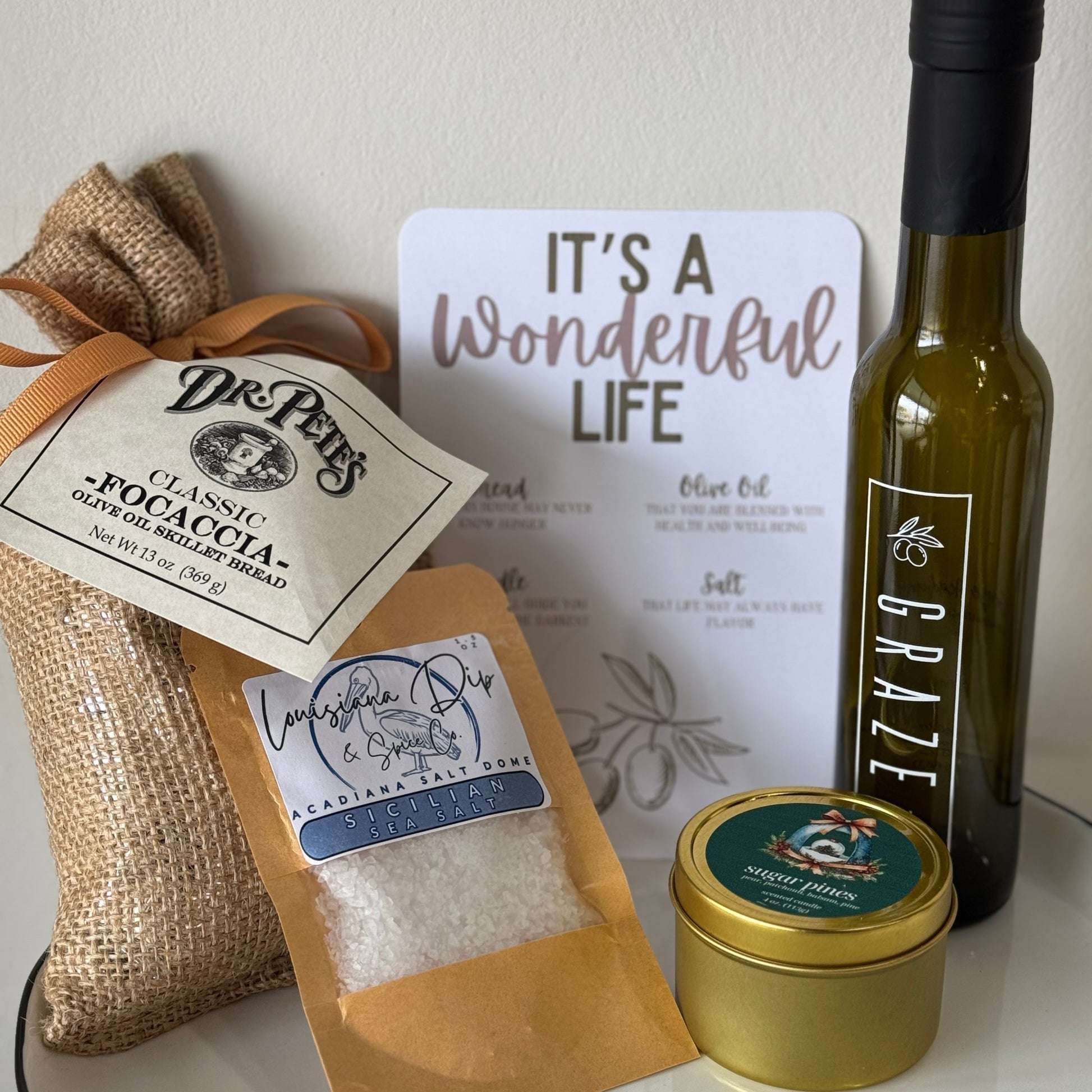 Exclusive Wonderful Life Collection of Dr. Petes bread, olive oil bottle, spice bag, a candle, and a card on a white surface