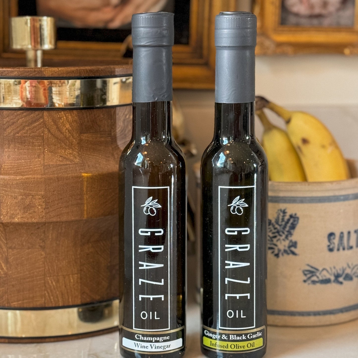 Two bottles of Graze Oil on a surface with a wooden barrel and bananas in the background.