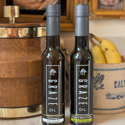 Two bottles of Graze Oil on a surface with a wooden barrel and bananas in the background.