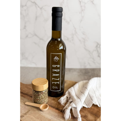 Bottle of Grazeo olive oil on a wooden board with a jar and cloth in the background.