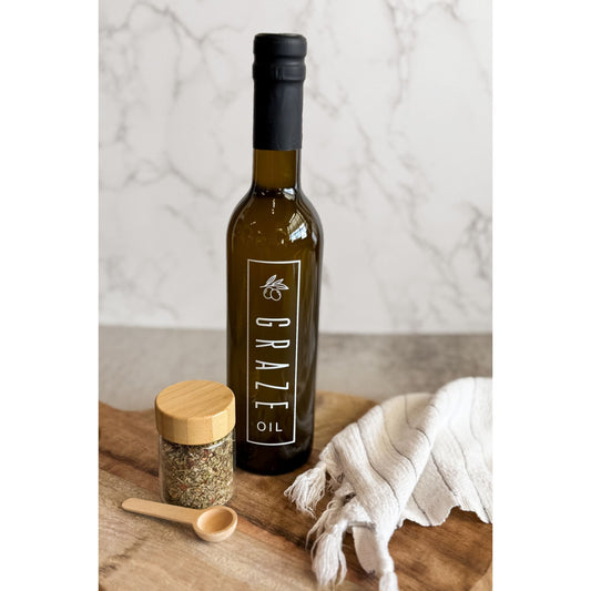 Bottle of Grazeo olive oil on a wooden board with a jar and cloth in the background.