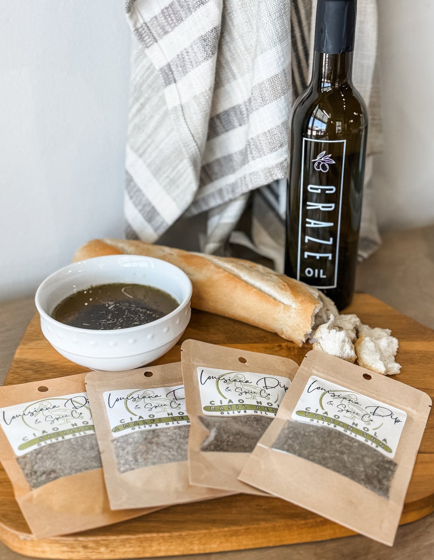 Bottle of Graze Oil olive oil, a bowl of oil, and dip packages with a baked baguette on a wooden tray with a towel in the background.