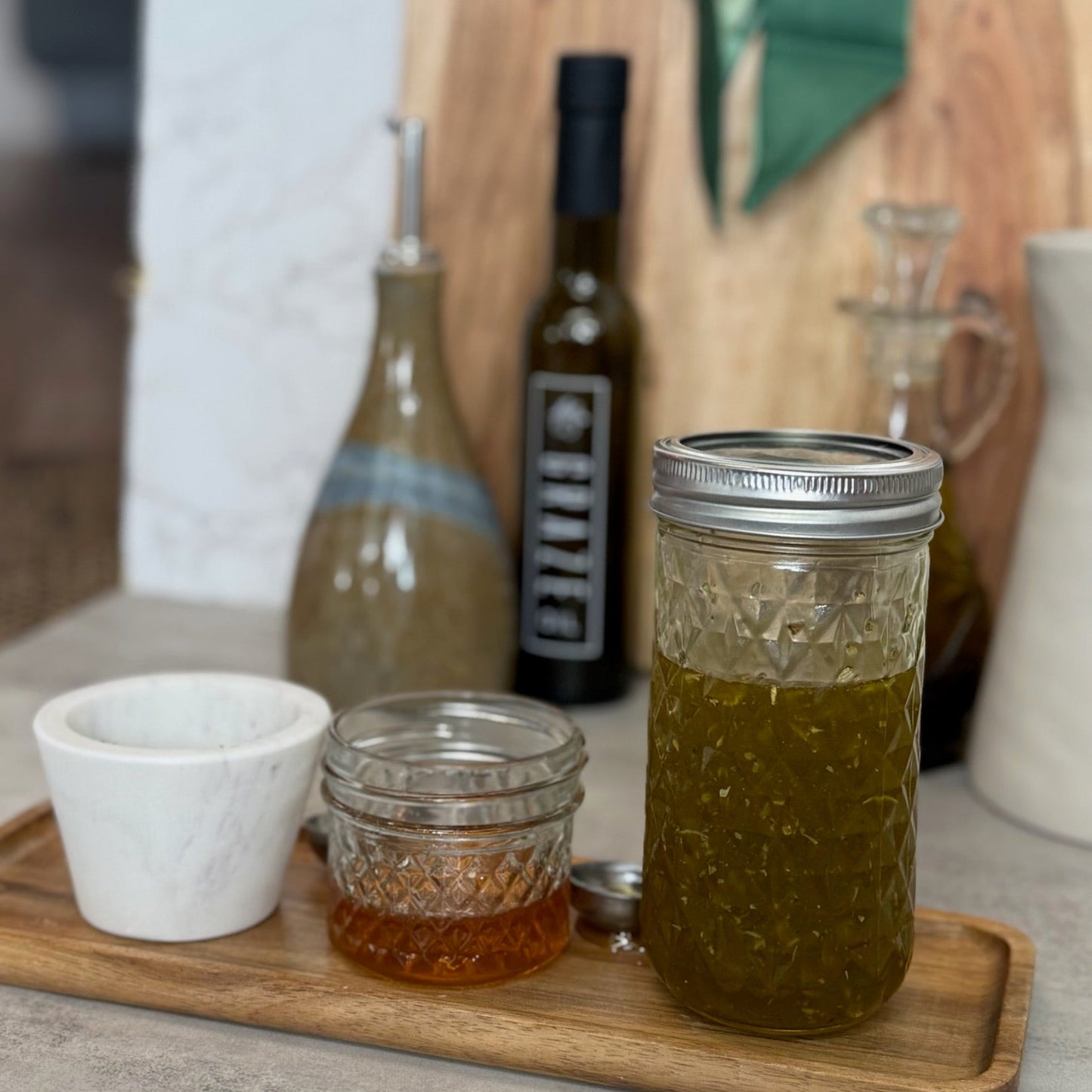 A glass mason jar filled with olive oil sits on a wooden tray next to a small white cup and a small glass jar of honey, with Graze Oil olive oil bottles and a wooden cutting board in the background.