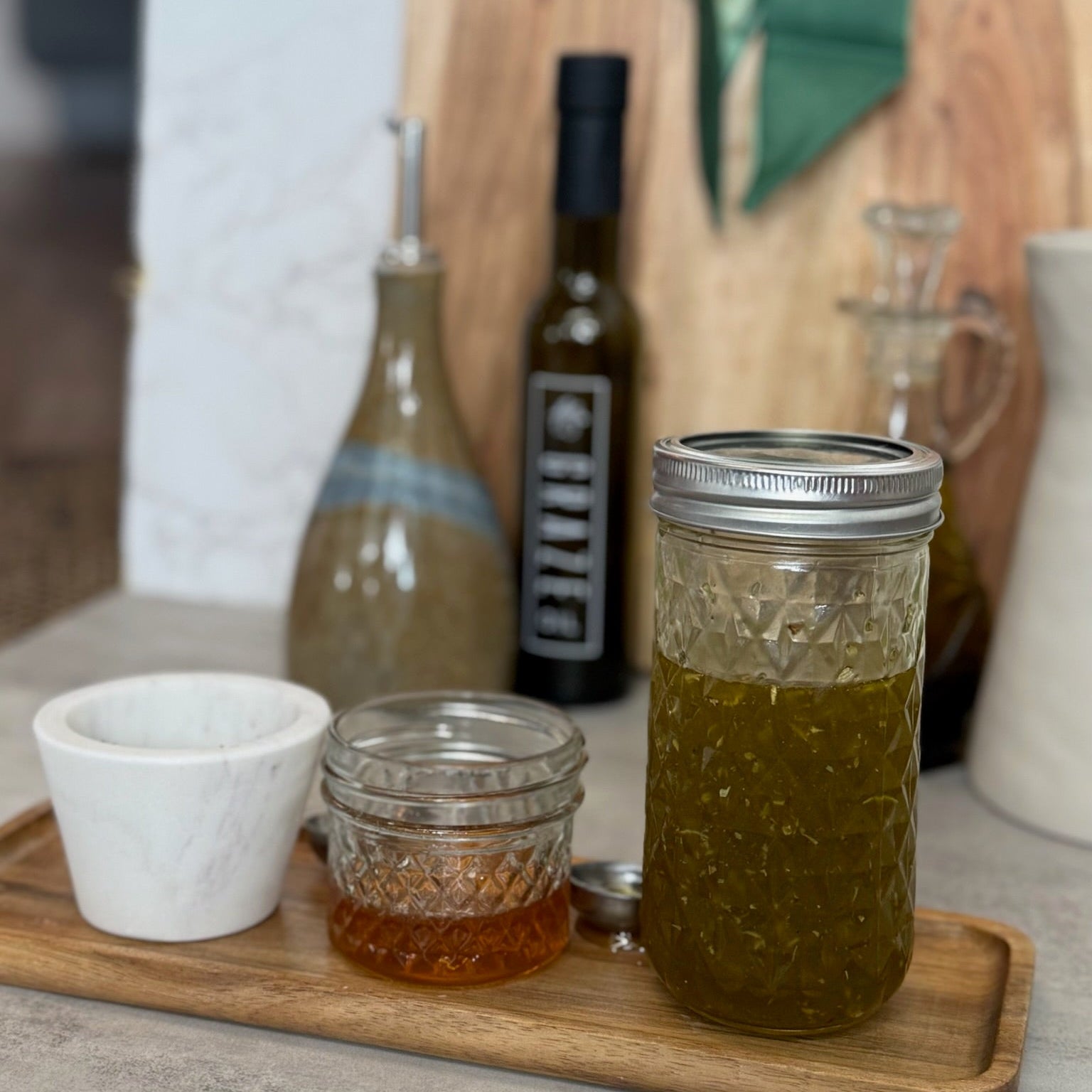 A glass mason jar filled with olive oil sits on a wooden tray next to a small white cup and a small glass jar of honey, with Graze Oil olive oil bottles and a wooden cutting board in the background.