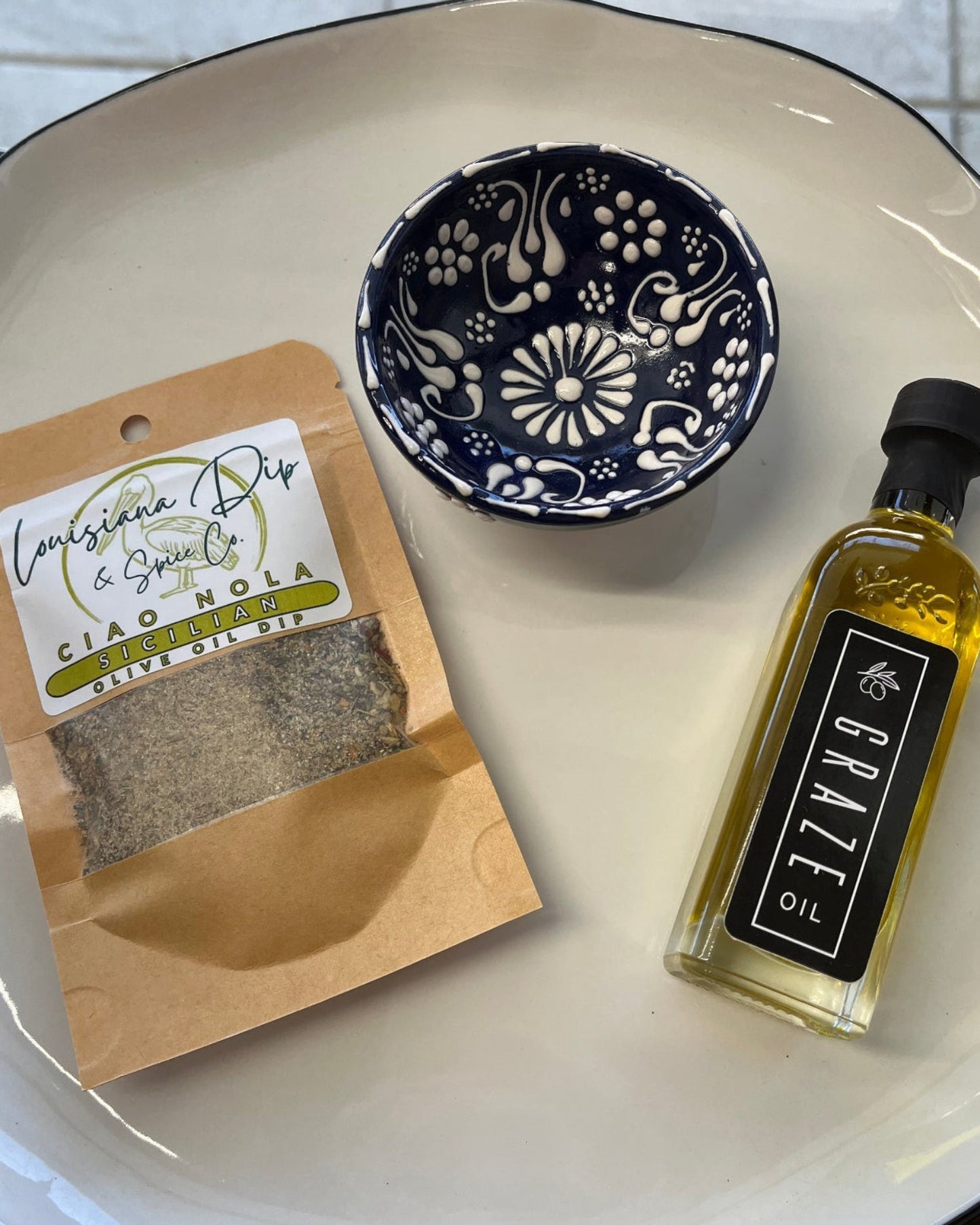 Tray with a bread dip package, small bowl, and a graze olive oil bottle on a tiled floor background