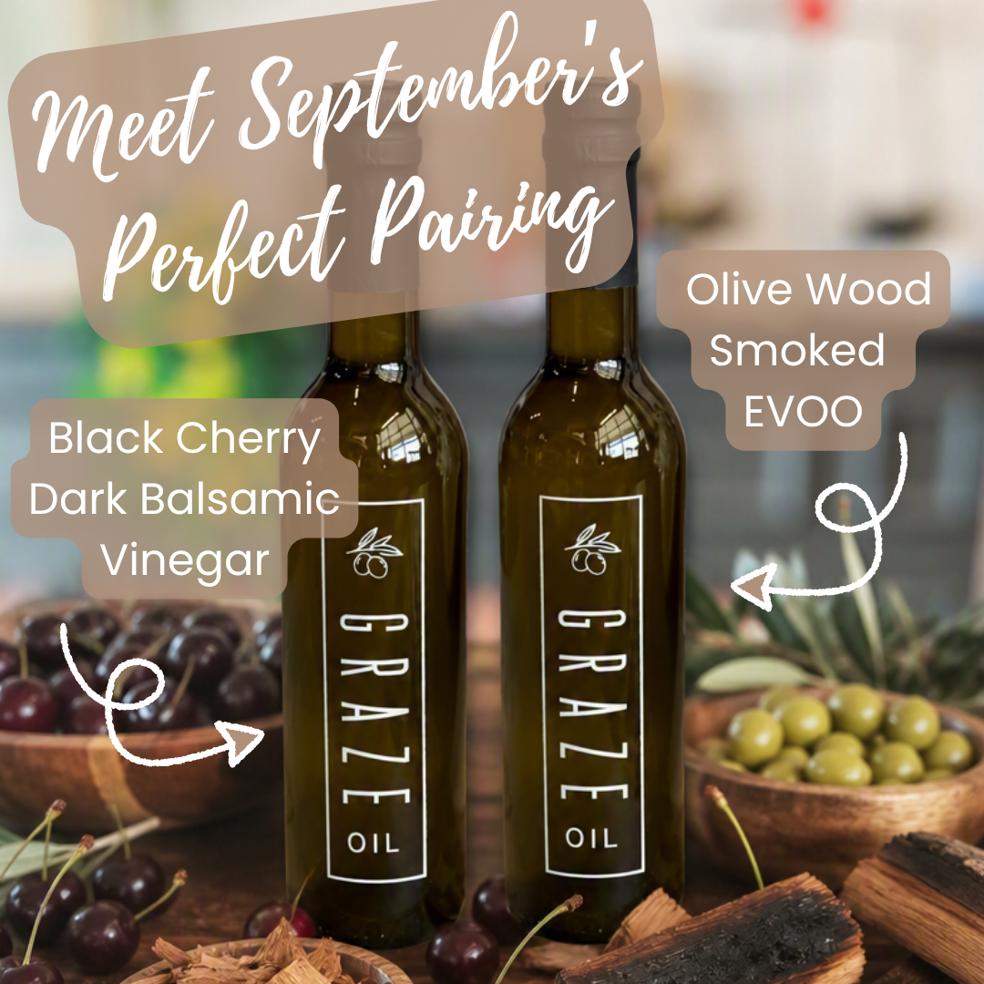Two dark glass bottles labeled GRAZE Oil stand on a rustic wooden surface with bowls of cherries, olives, bark, and handwritten callouts reading 'Black Cherry Dark Balsamic Vinegar' and 'Olive Wood Smoked EVOO' plus a banner that says 'Meet September's Perfect Pairing.'.