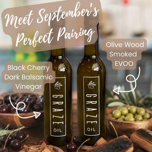 Two dark glass bottles labeled GRAZE Oil stand on a rustic wooden surface with bowls of cherries, olives, bark, and handwritten callouts reading 'Black Cherry Dark Balsamic Vinegar' and 'Olive Wood Smoked EVOO' plus a banner that says 'Meet September's Perfect Pairing.'.