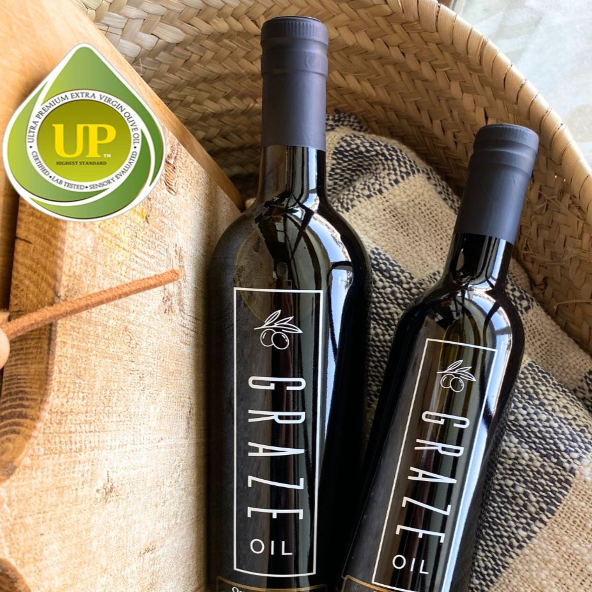 Two bottles of Ultra Premium Graze Oil on a woven basket with a checkered cloth underneath with UP tag