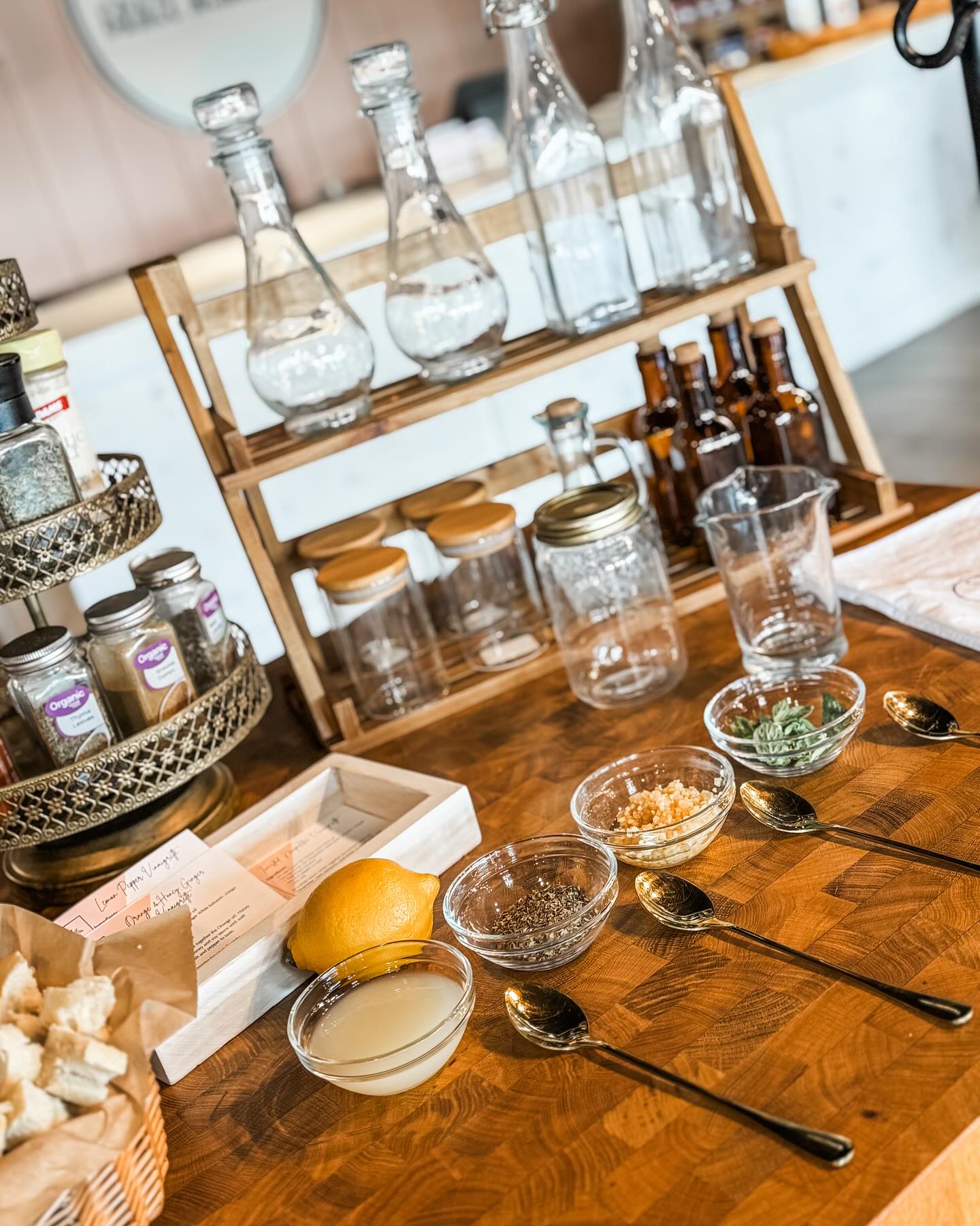 A rustic wooden table styled with vinaigrette ingredients, a lemon, small glass bowls with spices, metal spoons, glass bottles, Graze Oil and a wooden spice rack in a warm kitchen setting.