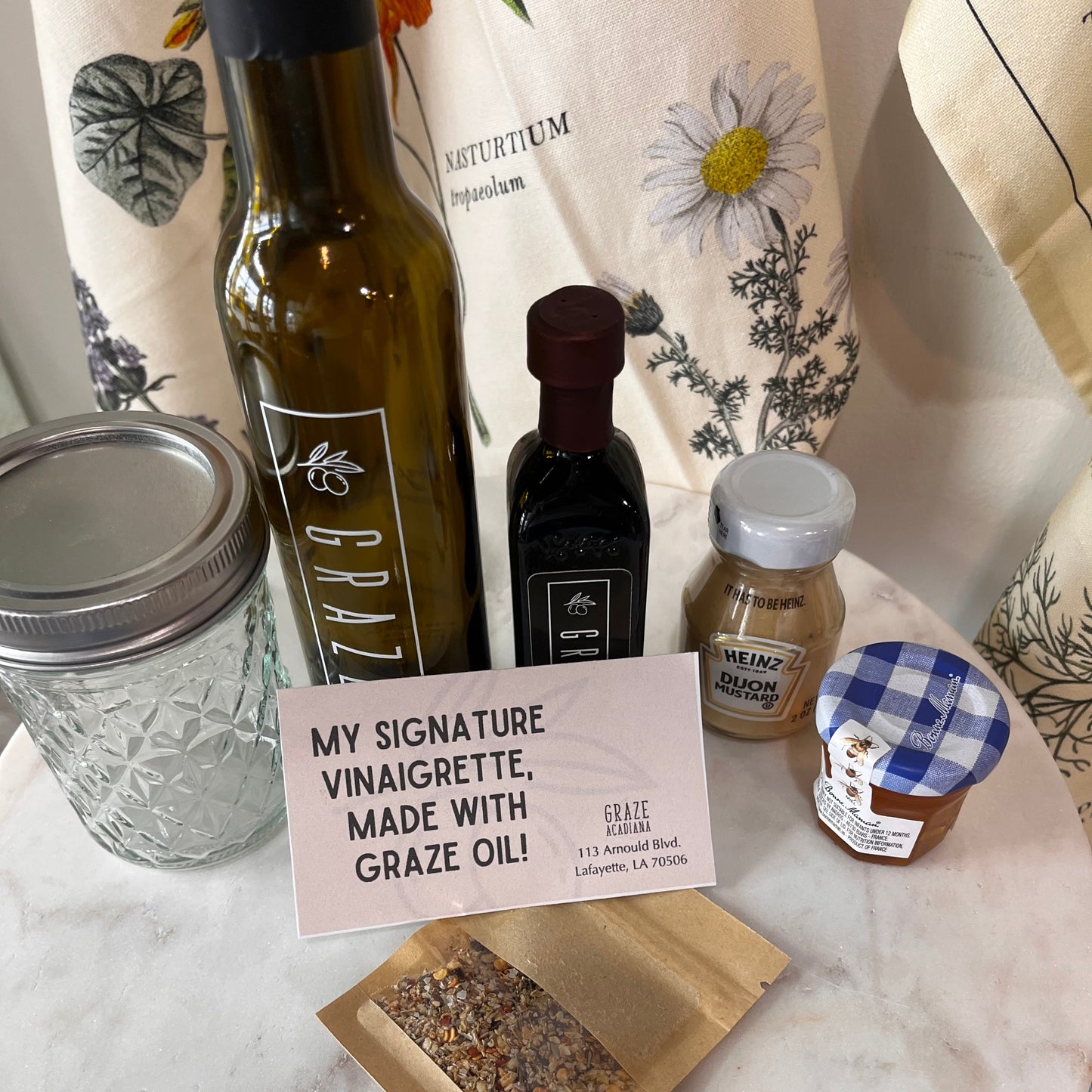A styled product display on a white marble surface featuring a bottle of Erazz grain-free vinaigrette, a small jar of organic apple butter with a blue gingham lid, a jar of honey, and a kraft pouch of seasoning mix, alongside a pink sign reading 'MY SIGNATURE VINAIGRETTE, MADE WITH GRAZE OIL!'.