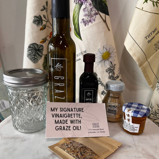 A tabletop display of gourmet food items including a bottle of Graze oil, a small dark bottle, a honey jar, a jarred product with a blue-and-white check lid, a mason jar, and a kraft pouch of seeds, arranged on a white marble surface with patterned fabric in the background. And a sign reading 'MY SIGNATURE VINAIGRETTE, MADE WITH GRAZE OIL!'.