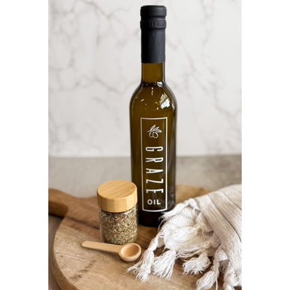 Bottle of Graze Oil on a wooden cutting board with a jar of spices and a white cloth.
